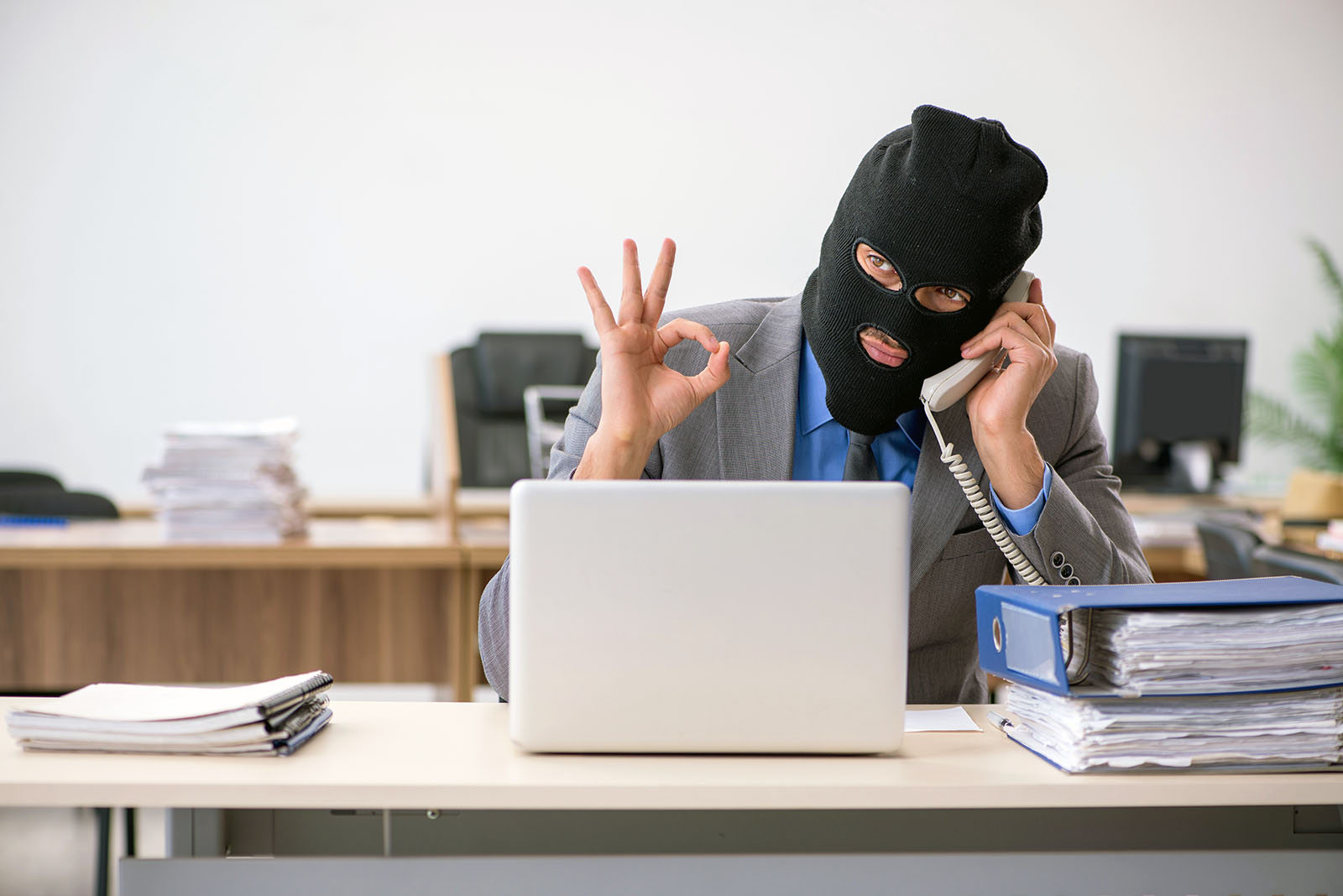 man in business suit and black ski mask flashing OK sign while sitting at desk with phone to ear