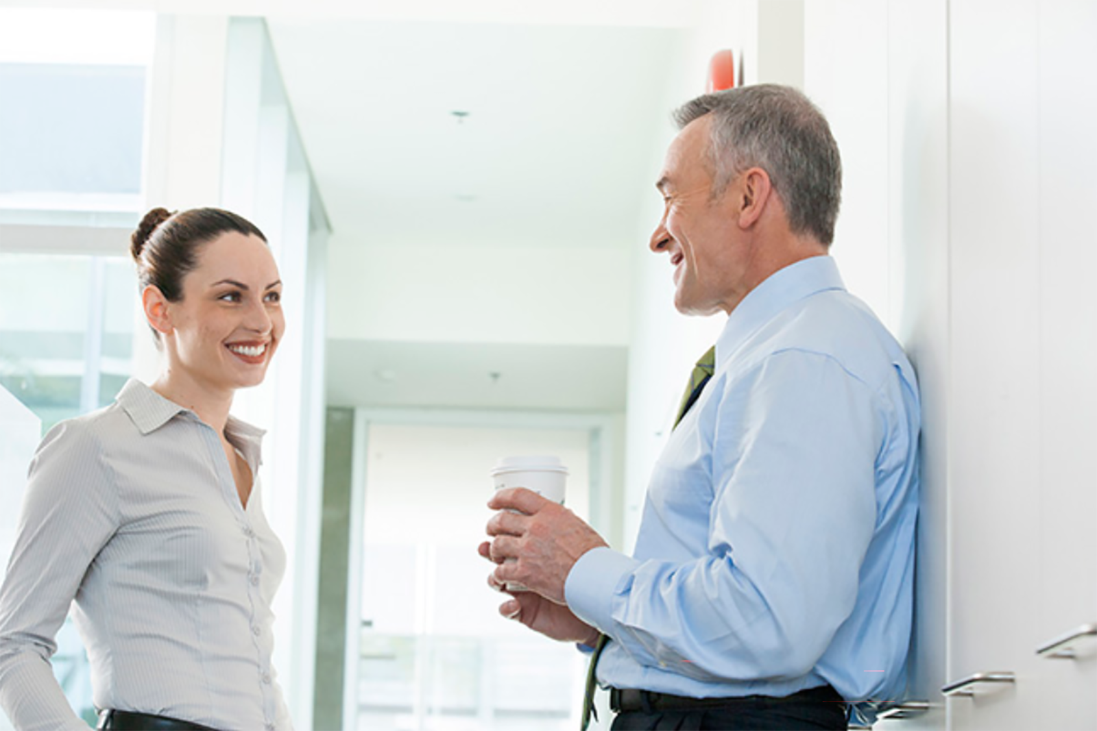 Younger businesswoman and older businessman smiling and talking in office hallway